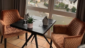Een balkon met twee comfortabele stoelen, een klein tafeltje en een bloempot. Een raam biedt uitzicht op een stad met een kerk en huizen.