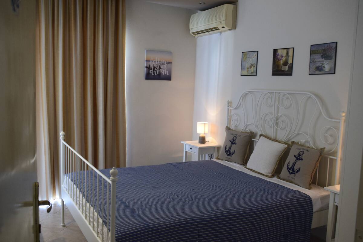 A bedroom with a bed, a nightstand, a lamp, and a wall with framed pictures.