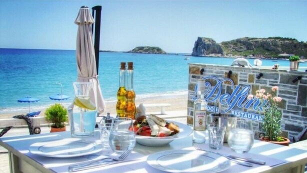 A table set for two with a view of the sea, a stone wall with a restaurant sign, and a bottle of liquor. - Delfini Beach Hotel (Rhodes)