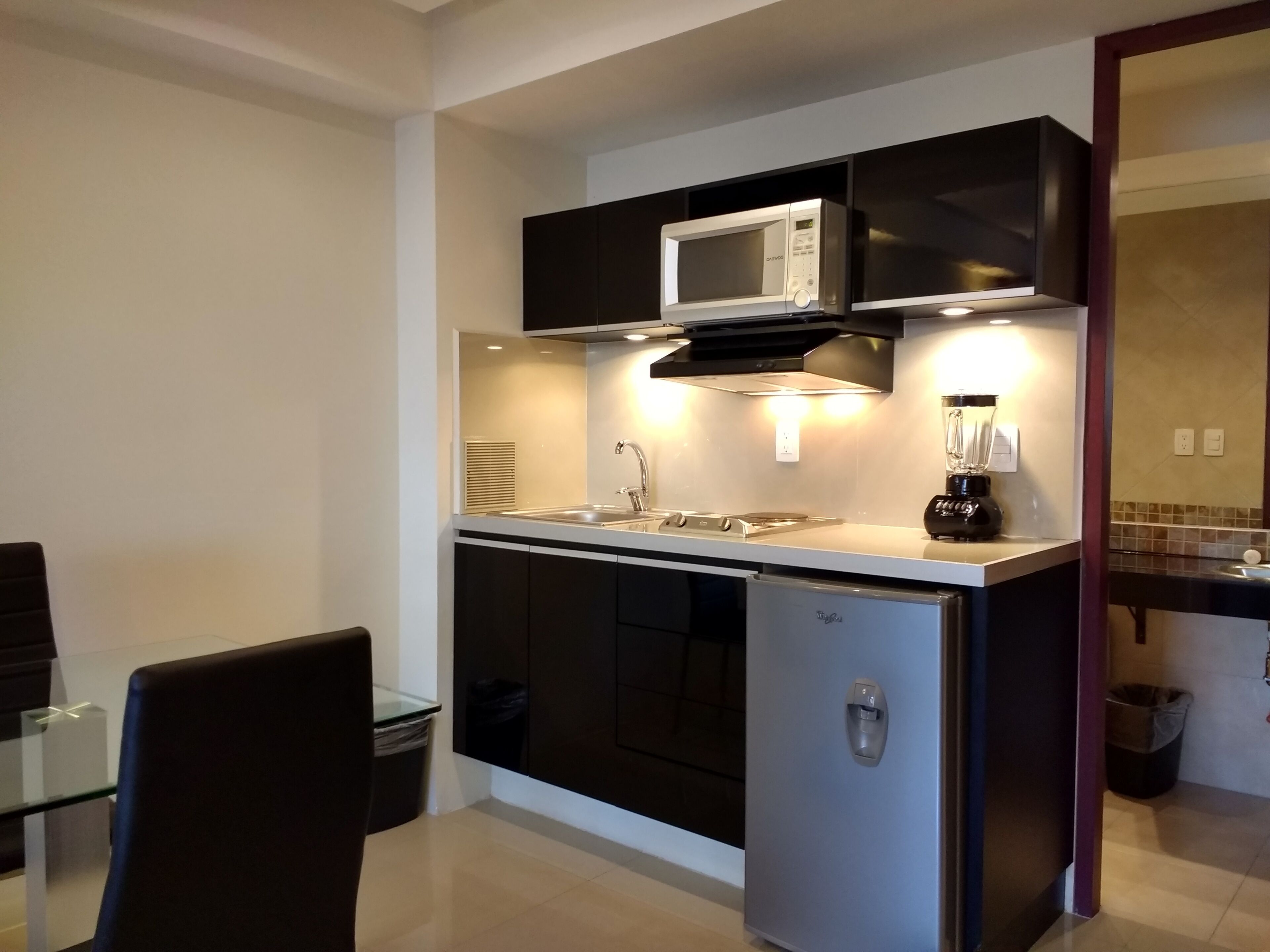 A compact kitchen with a microwave, stove, and sink. There is a trash bin and a glass table with chairs.