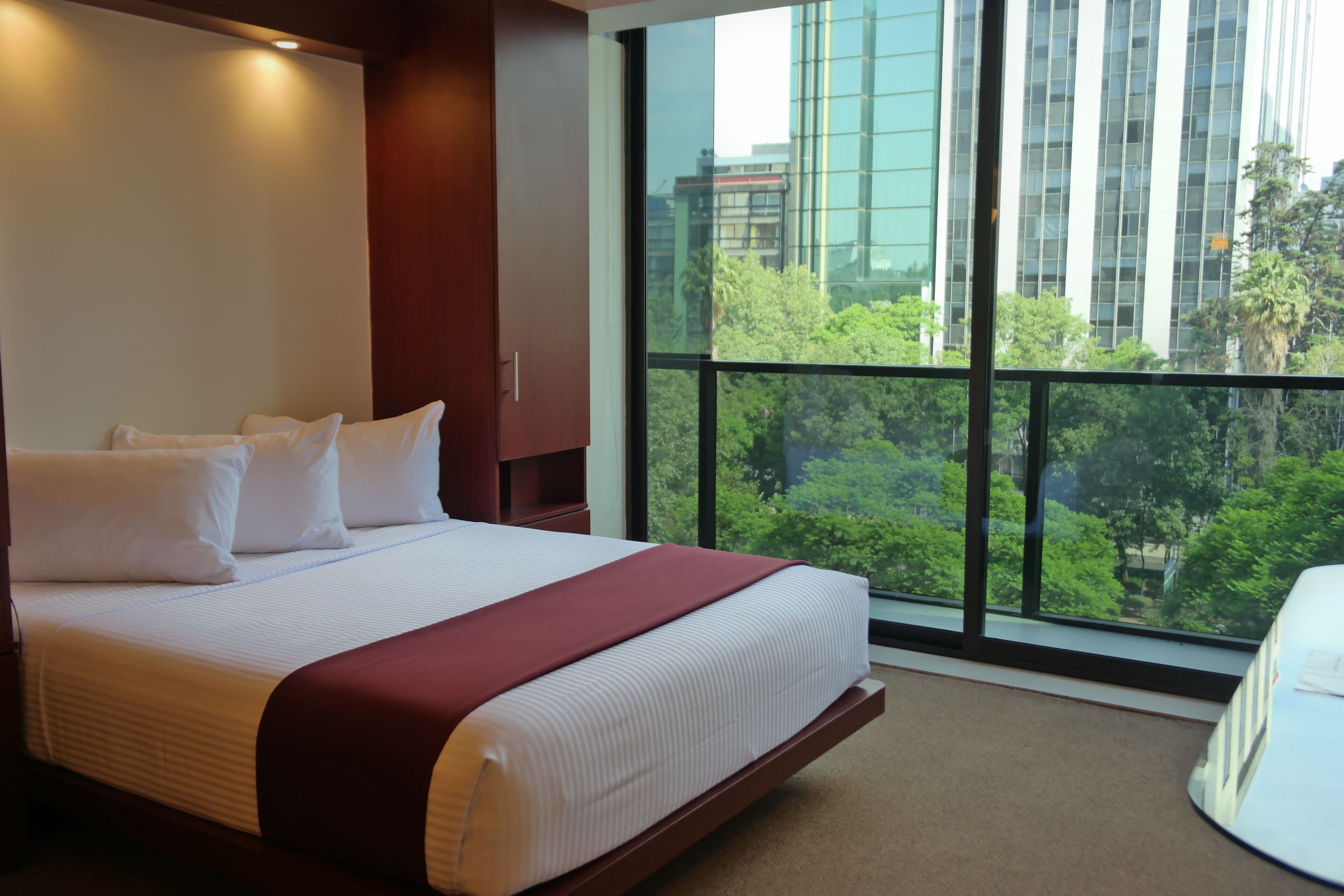 A hotel room with a large bed, a view of the city through a large window, and a balcony with a view of trees and buildings.