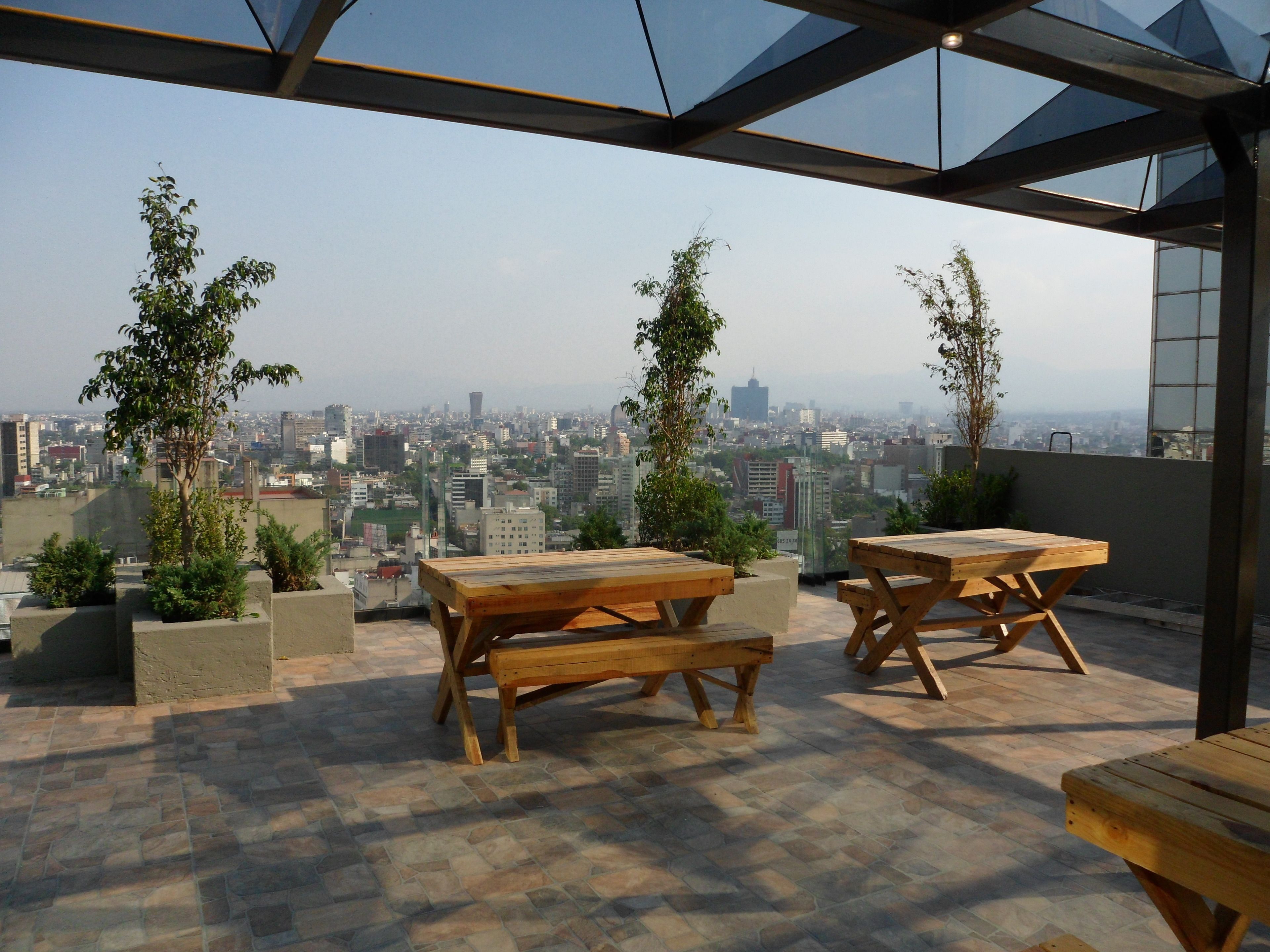 Terraço no telhado com mesas e bancos de madeira, plantas em vasos e vista para a cidade.