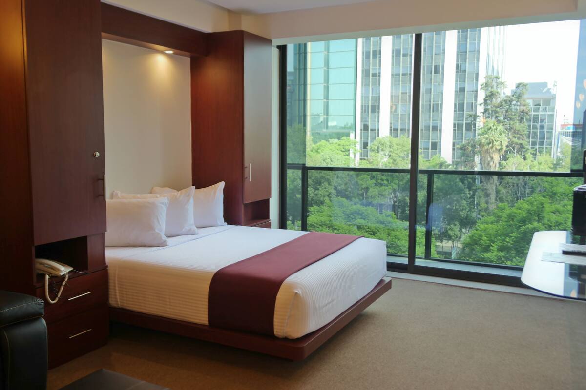 Ein Hotelzimmer mit einem großen Bett, einem Schreibtisch und Blick auf die Stadt durch ein großes Fenster.