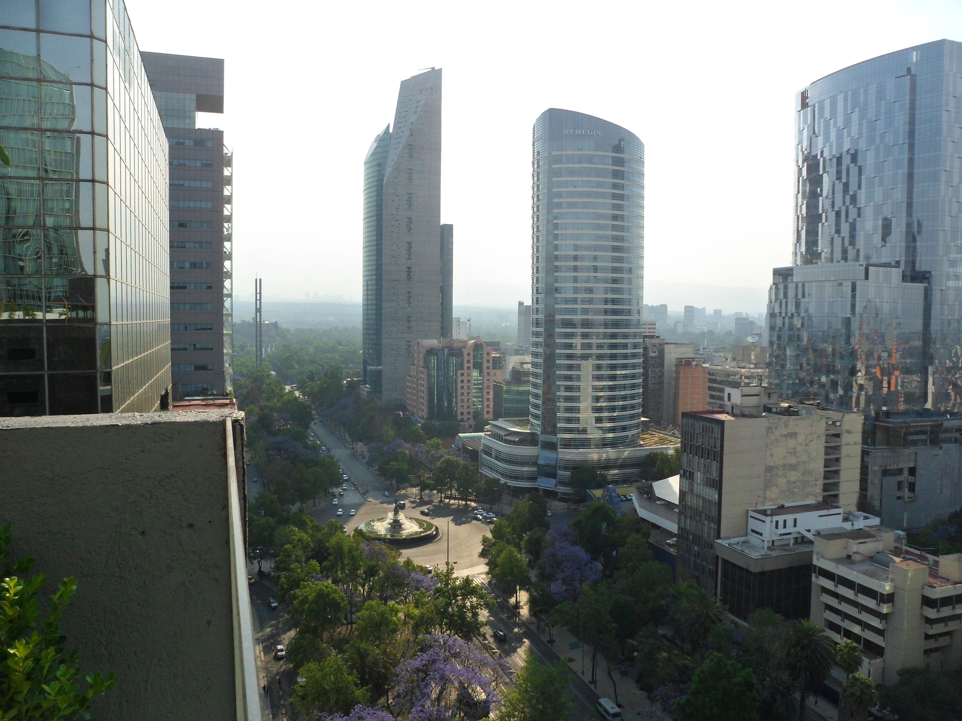 Un paisaje urbano con edificios modernos altos, una rotonda central y zonas verdes circundantes.