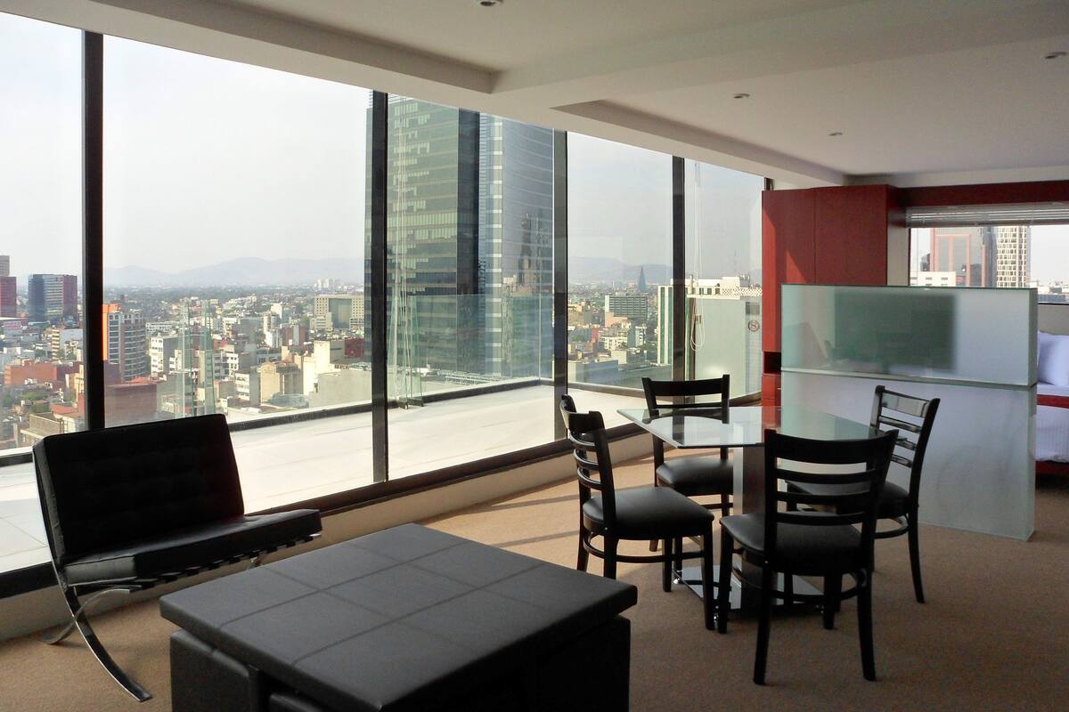 Ein modernes Hotelzimmer mit großem Fenster und Blick auf die Stadt, einem Essbereich mit Tisch und Stühlen sowie einem an der Wand befestigten Flachbildfernseher.