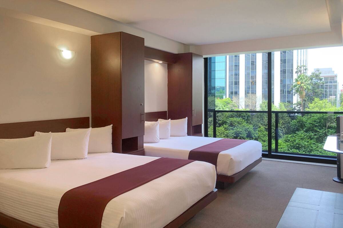 Ein Hotelzimmer mit zwei Betten, einem großen Fenster und Blick auf Gebäude und Grünflächen, sowie einem kleinen Tisch.