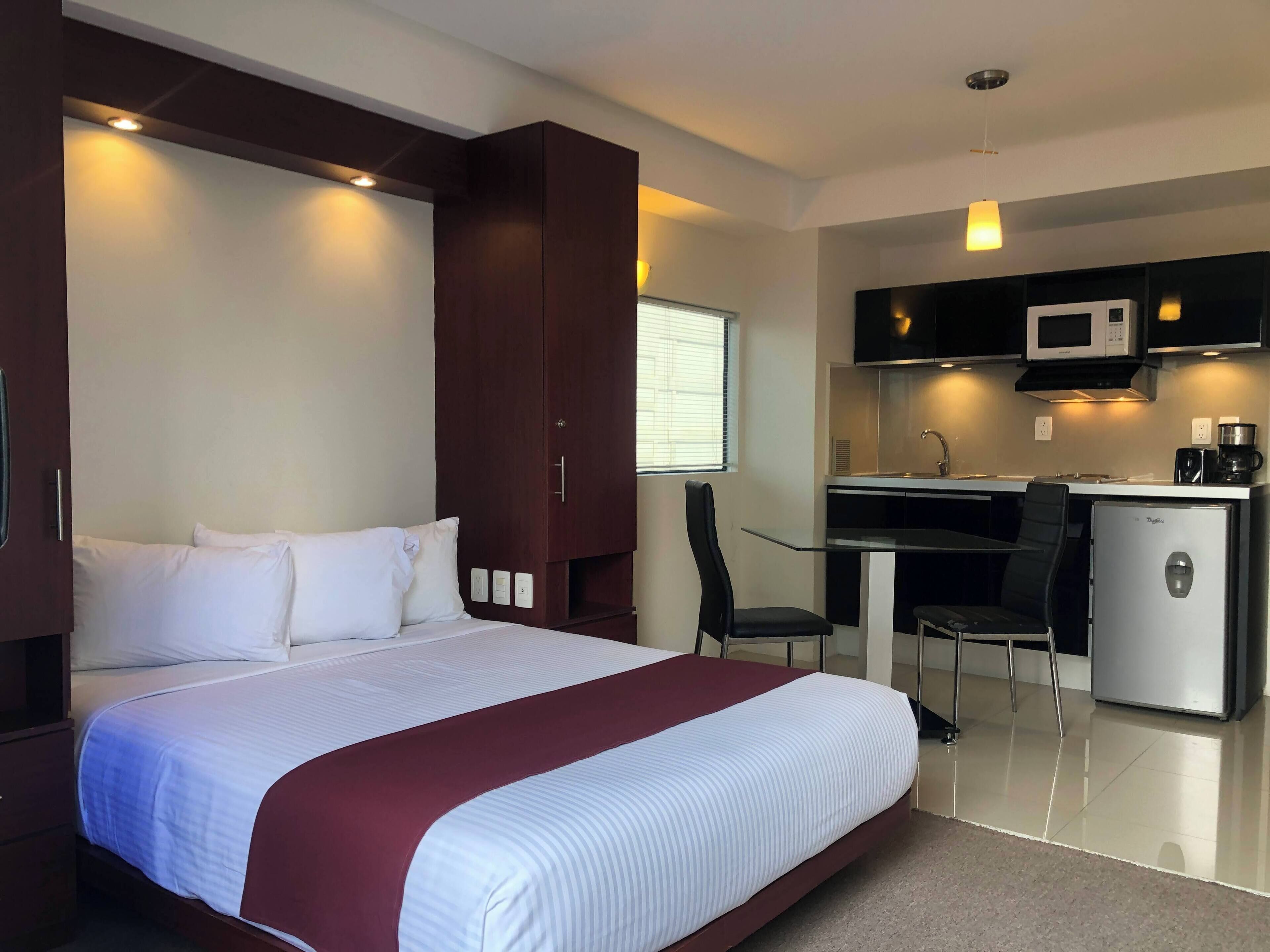 Habitación de hotel con cama, escritorio, silla, microondas, refrigerador y ventana con persianas.