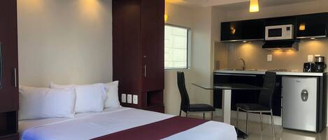 Habitación de hotel con cama, escritorio, silla, microondas, refrigerador y ventana con persianas.