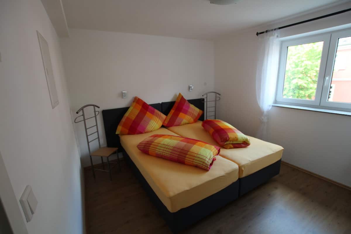 Quarto pequeno com uma cama de solteiro, piso de madeira, janela com vista para o verde e uma escada de metal encostada na parede.