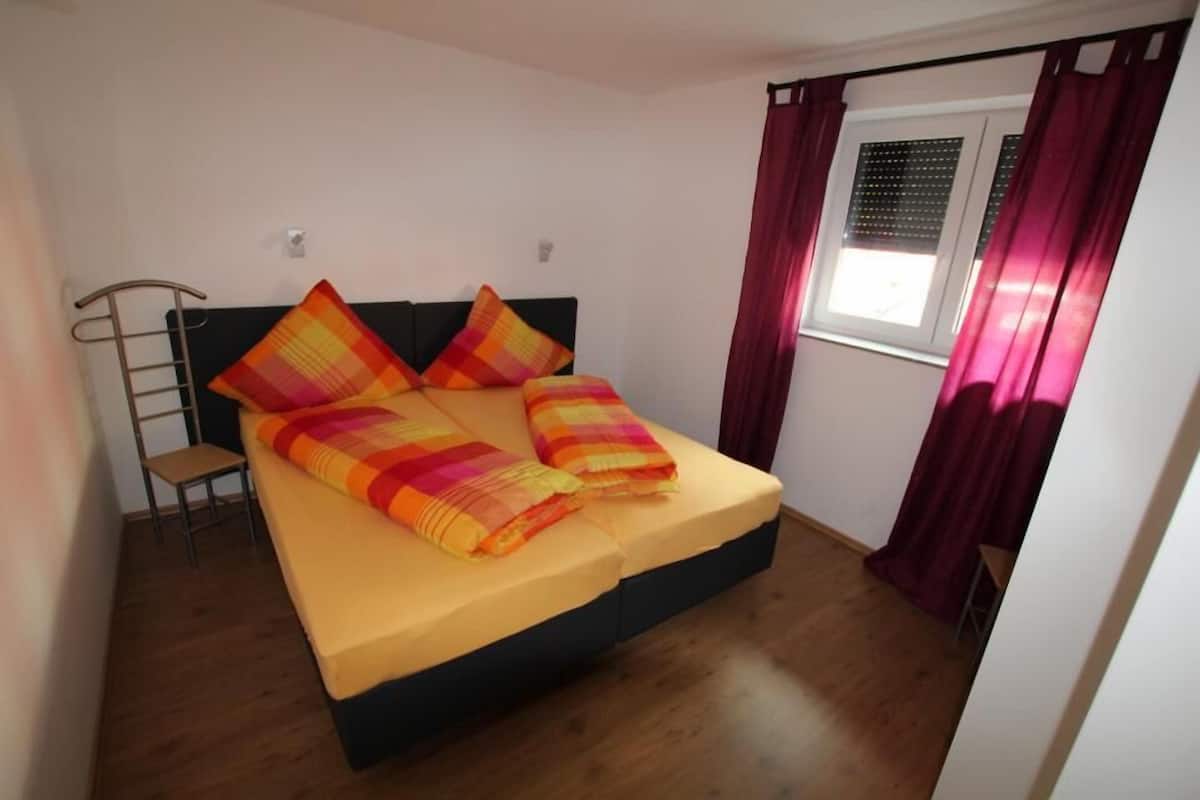 Uma cama com colchão amarelo e estrutura preta, duas almofadas, piso de madeira, uma cadeira e uma janela com cortinas.