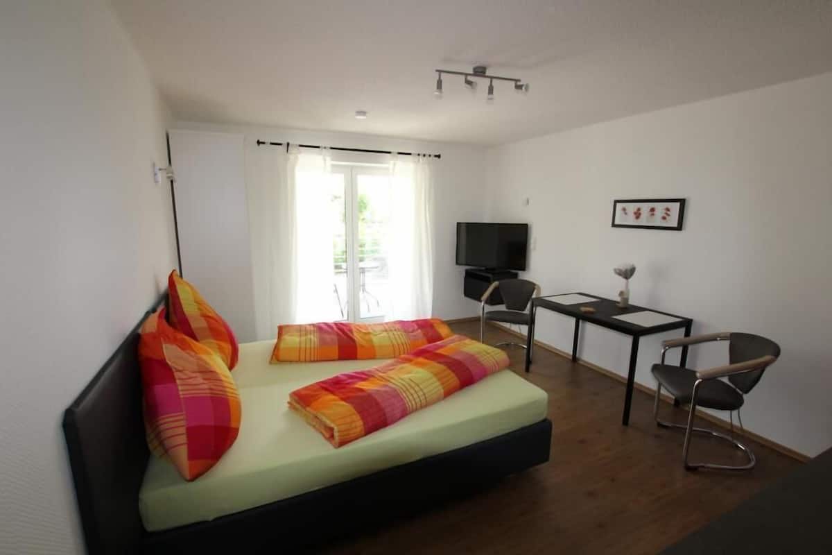 Quarto com cama, escrivaninha, cadeira, televisão e janela com cortinas.