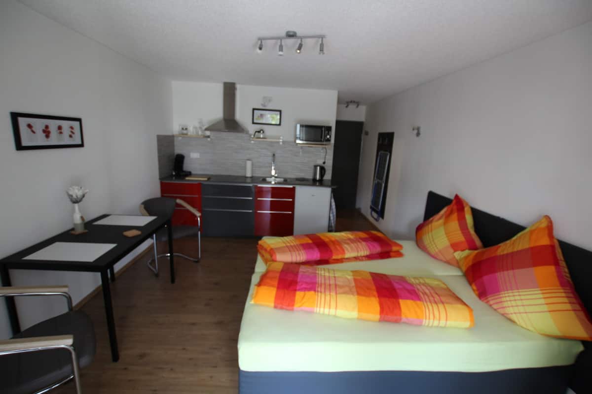 Quarto de hotel compacto com cama, escrivaninha, cadeira, cozinha compacta e pequena mesa de jantar.