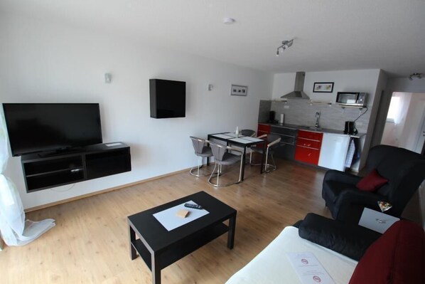 A modern living room with a flat-screen TV, a dining area with a table and chairs, a black sofa, and a kitchen area with a microwave and red cabinets.