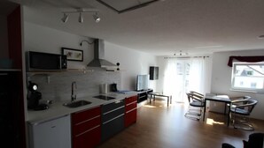 A modern kitchen with a microwave, sink, and red cabinets. There is a dining area with a table and chairs, and a window with curtains. - Apartment Wesseling Zentrum (Wesseling)