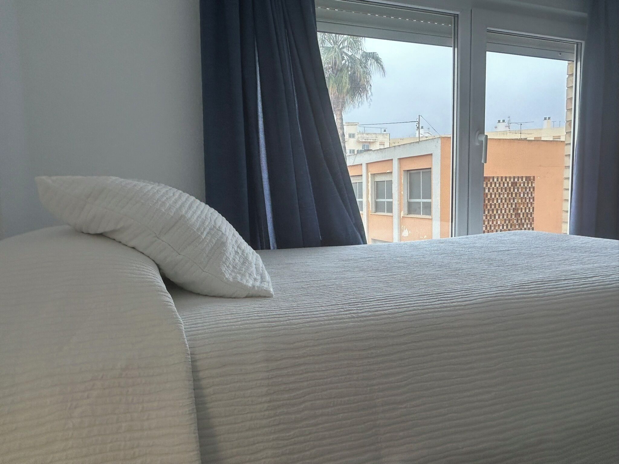 Ein Bett mit weißem Kopfkissen und grauer Decke, ein Fenster mit Blick auf Gebäude und eine Palme draußen.