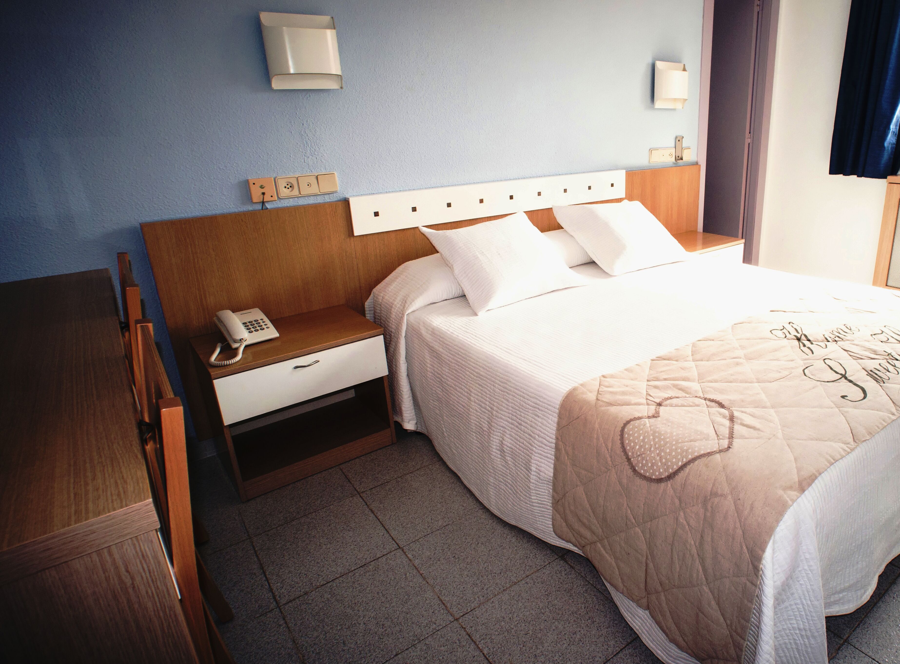 A hotel room with a wooden headboard, a bedside telephone, a white drawer, a bed with white bedding and a beige quilt, and two wall-mounted light fixtures.