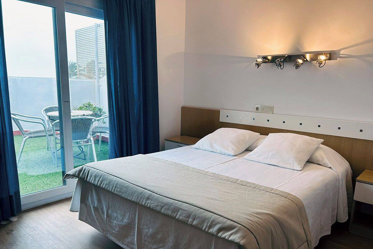 Habitación de hotel con cama, mesita de noche, lámpara de pared y vista a un espacio exterior con sillas y una mesa.
