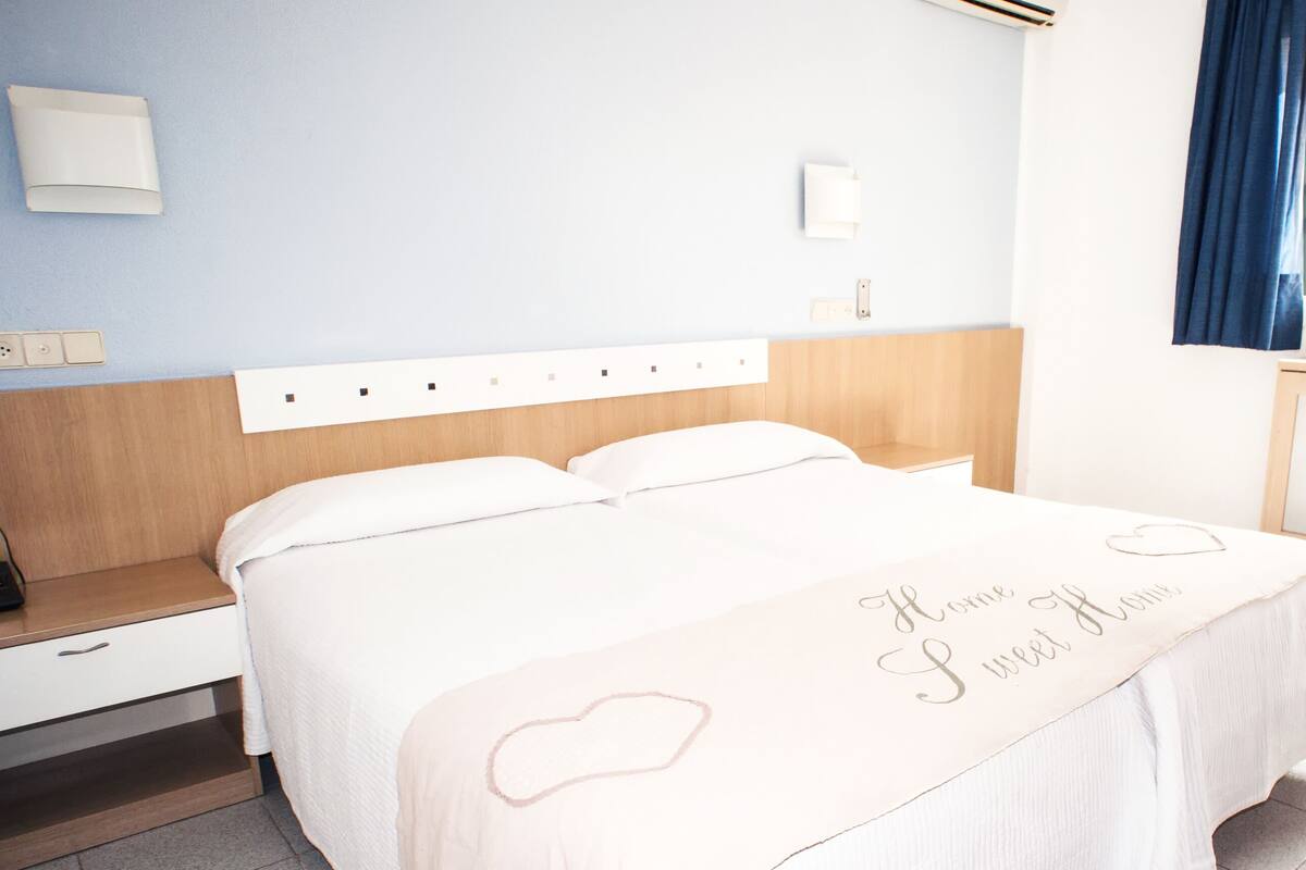 Habitación de hotel ordenada con cama, mesita de noche y ventana con cortinas.