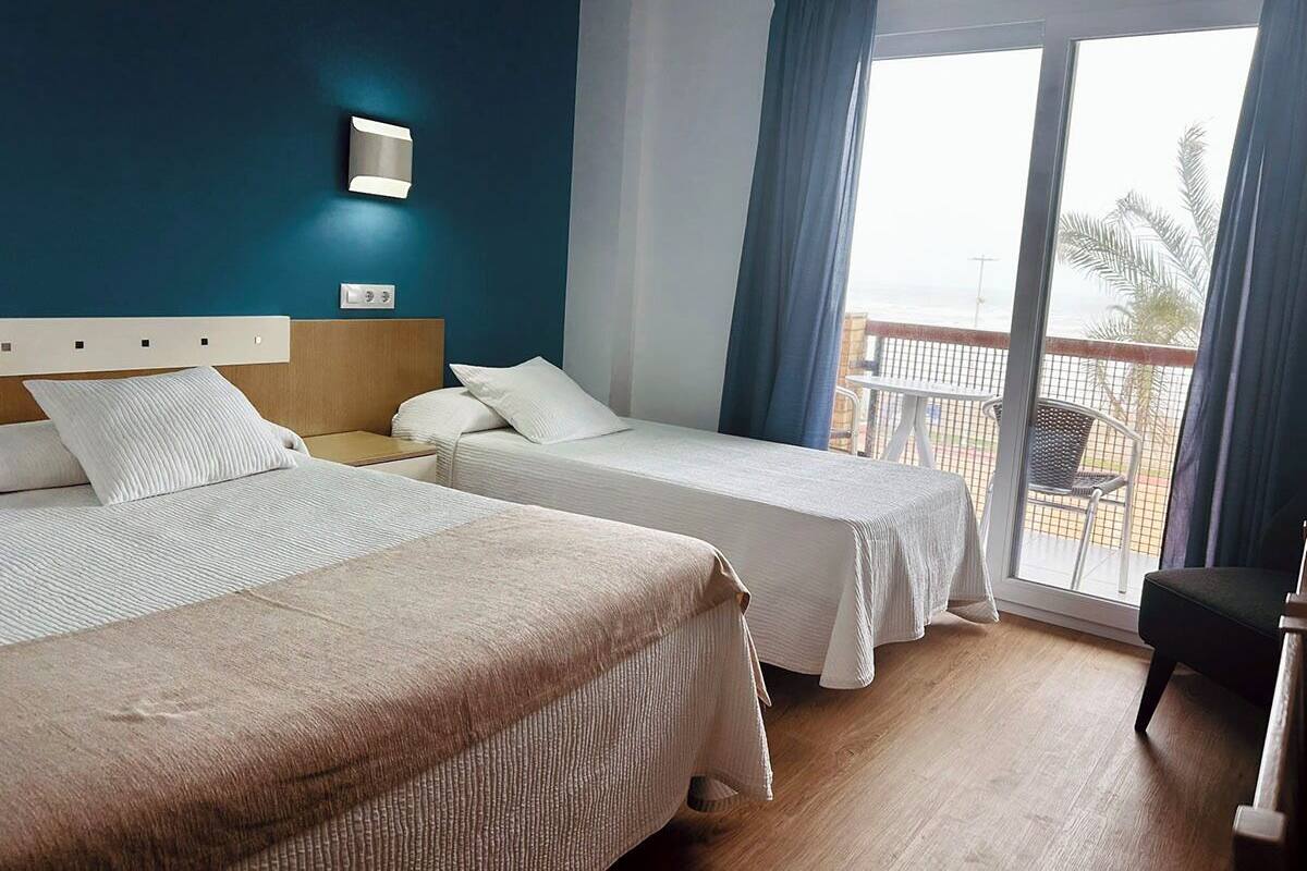 Habitación de hotel con dos camas, una mesita de noche, una lámpara de pared y un balcón con una silla.