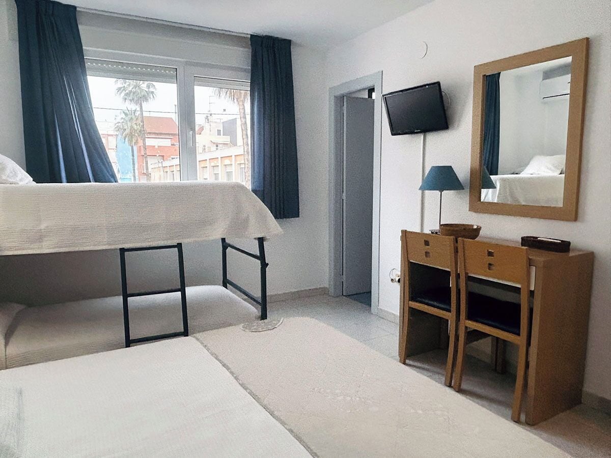 Quarto de hotel com cama, escrivaninha, cadeira, televisão e janela com cortinas.