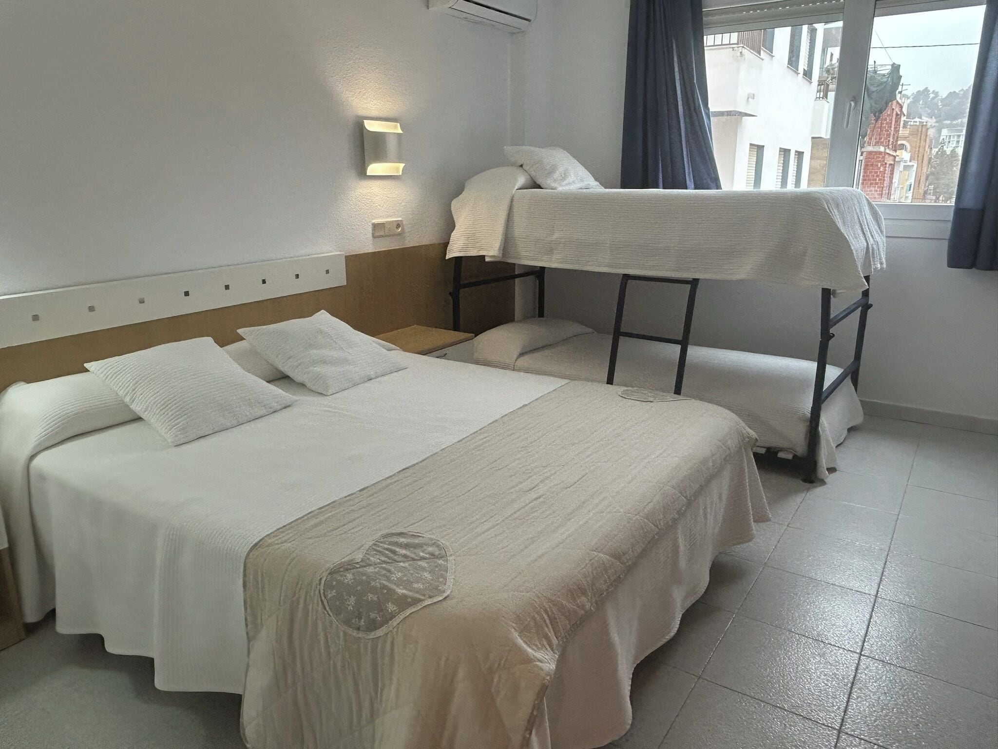 Hotellihuone, jossa on kaksi sänkyä, kerrossänky, ikkuna, jossa on verhot, ja seinään kiinnitetty valaisin.