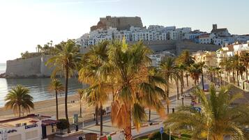 Un paisaje urbano costero con una estructura imponente similar a una fortaleza, palmeras bordeando el paseo marítimo y una vista despejada del mar.