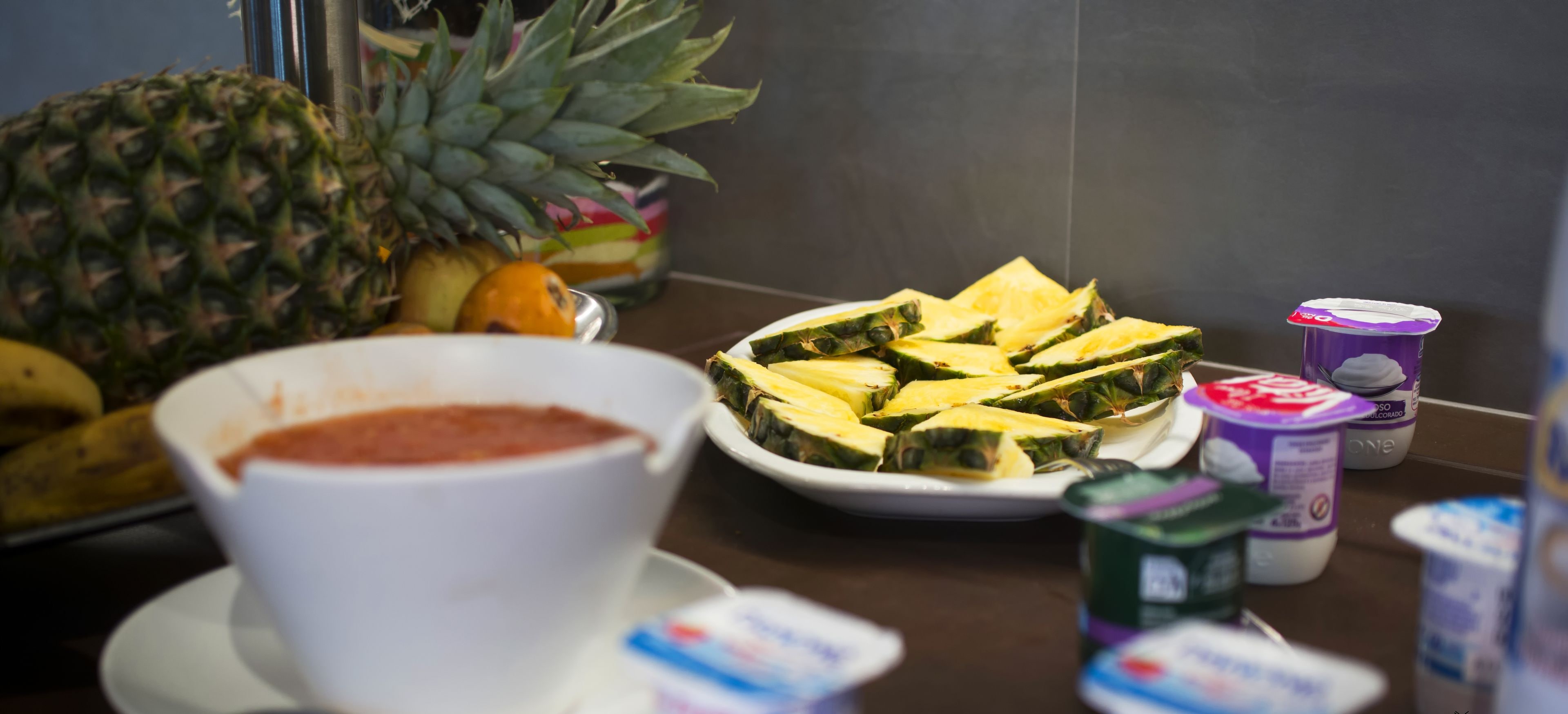 A kitchen counter with a bowl of soup, a plate of sliced fruit, and several containers of yogurt.