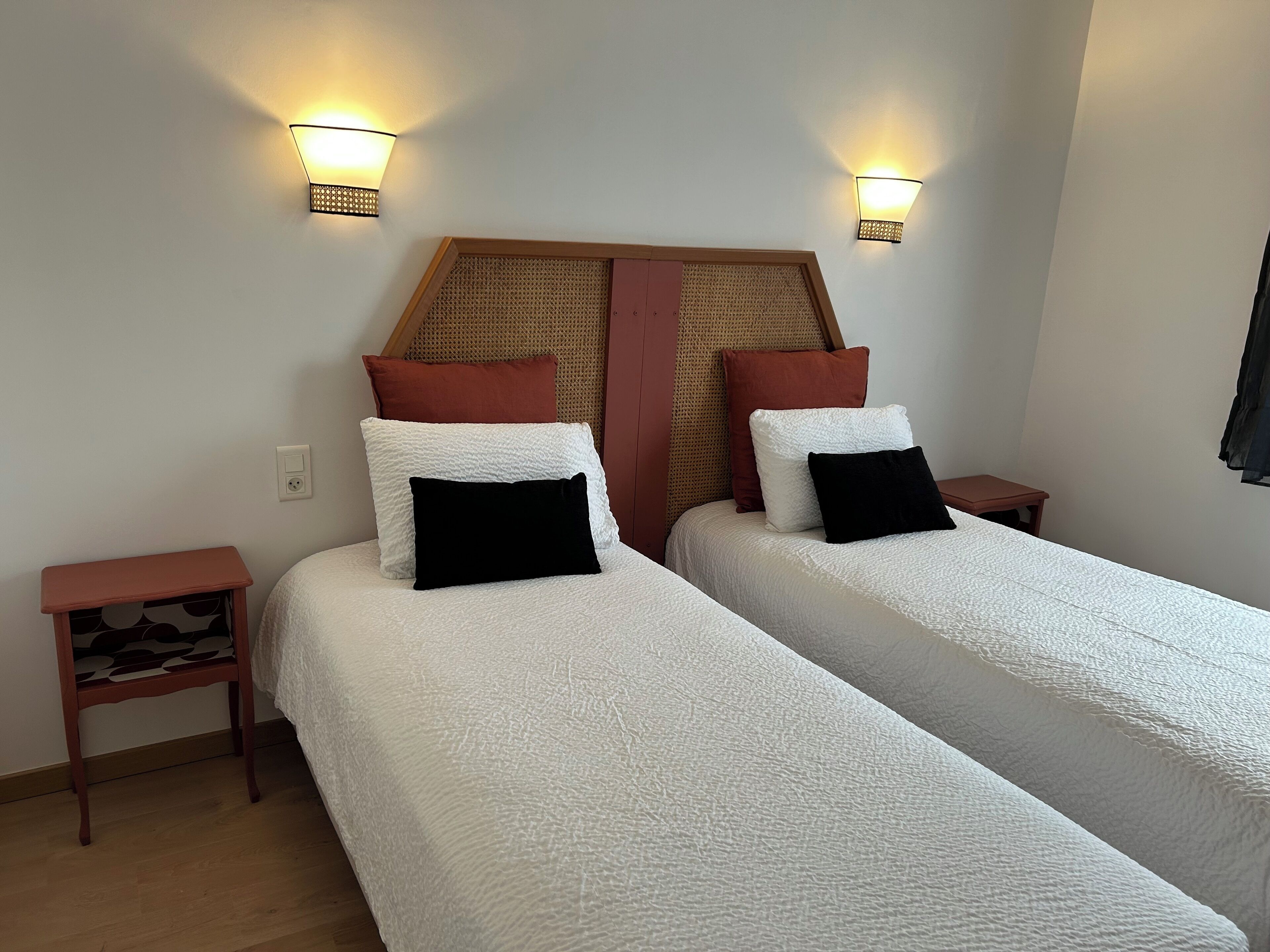 Cama doble con ropa de cama blanca y almohadas negras, dos mesitas de noche y luces fijadas a la pared.