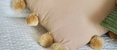 Un cojín color beige con detalles de pompones sobre una colcha blanca con textura.