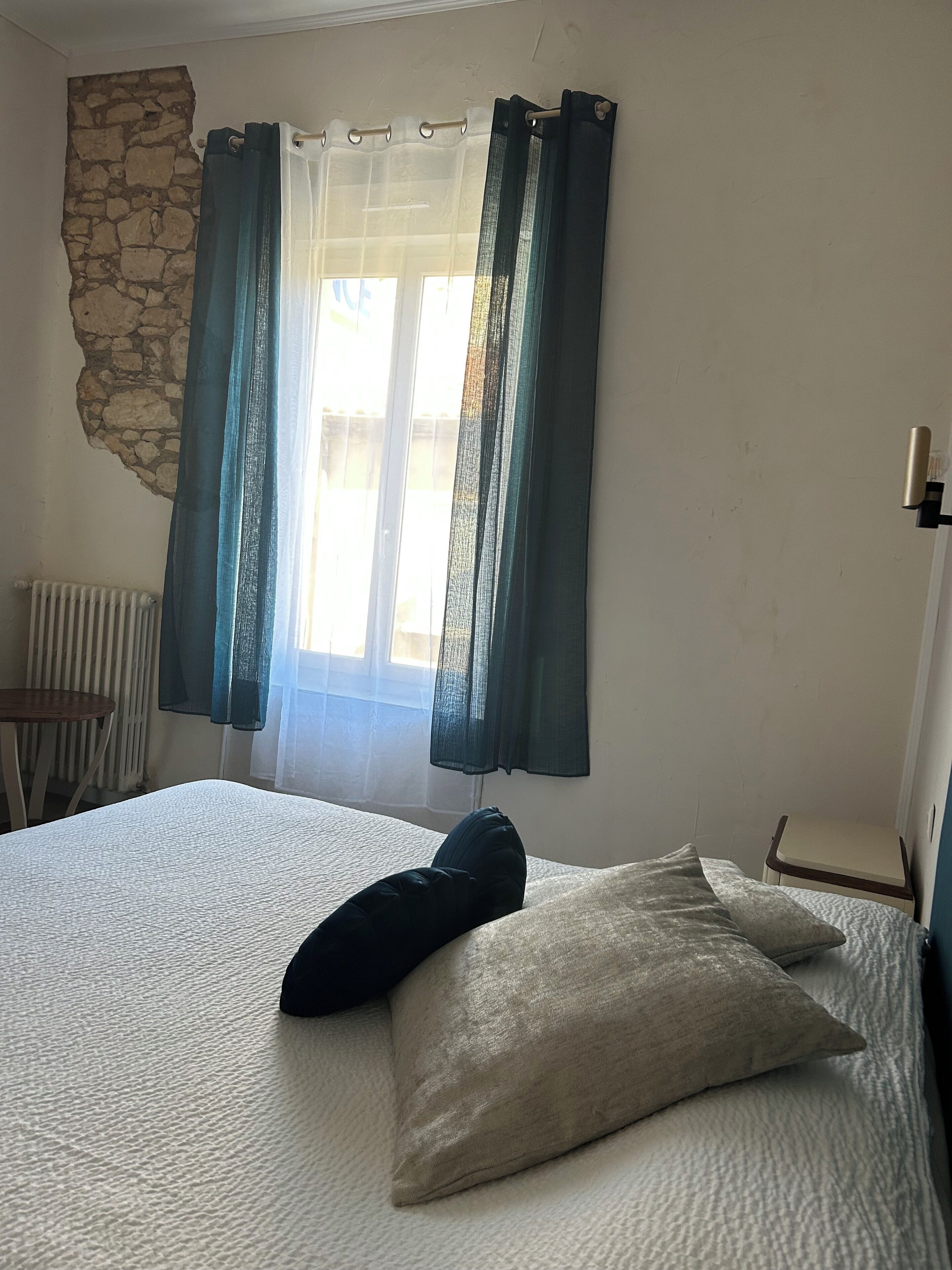 Une chambre à coucher avec un lit, un mur en pierre, un radiateur, une petite table et une fenêtre avec des rideaux.