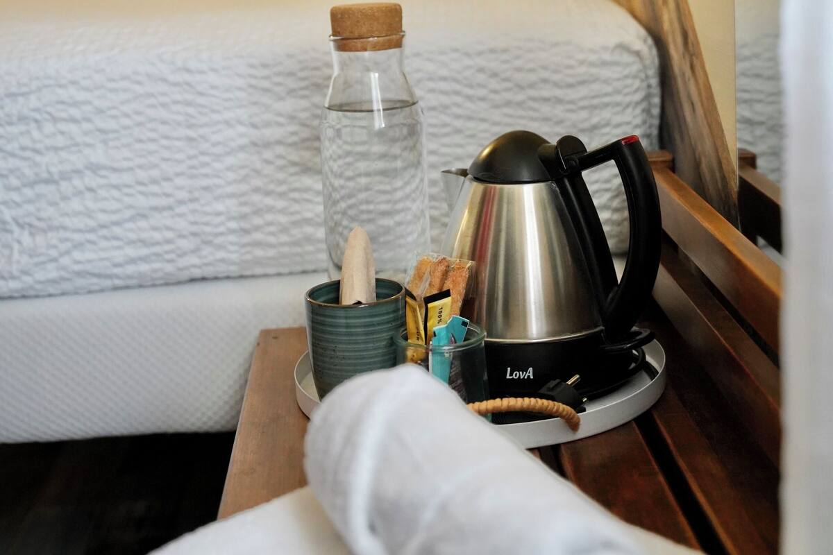 A neatly made bed with a pillow labeled "DU LARGE" and a wooden headboard. A small table beside the bed holds a glass bottle, a kettle, and a cup.
