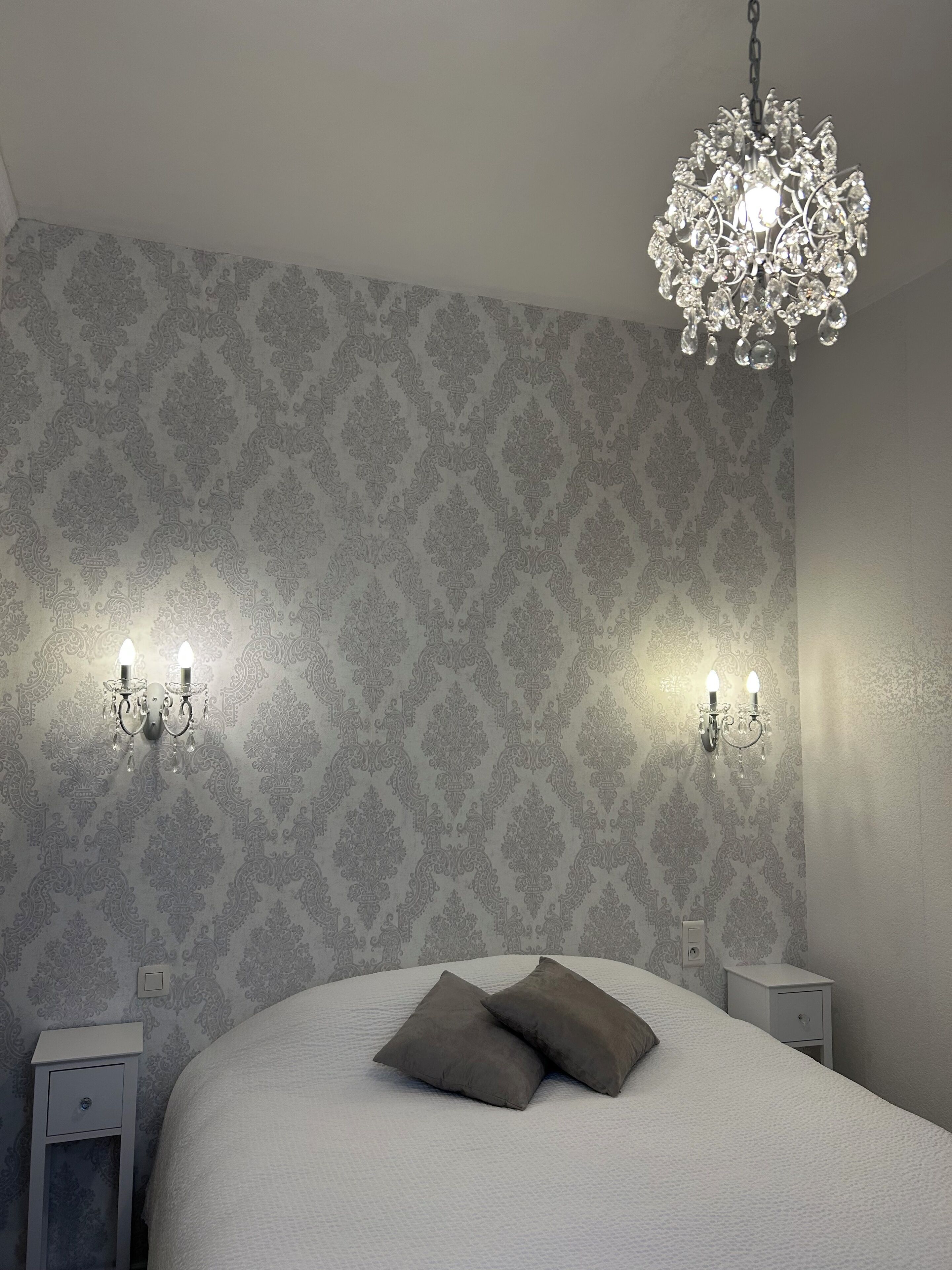 Quarto com papel de parede estampado, um candelabro, duas arandelas, uma cama com almofadas e uma mesa de cabeceira com gaveta.