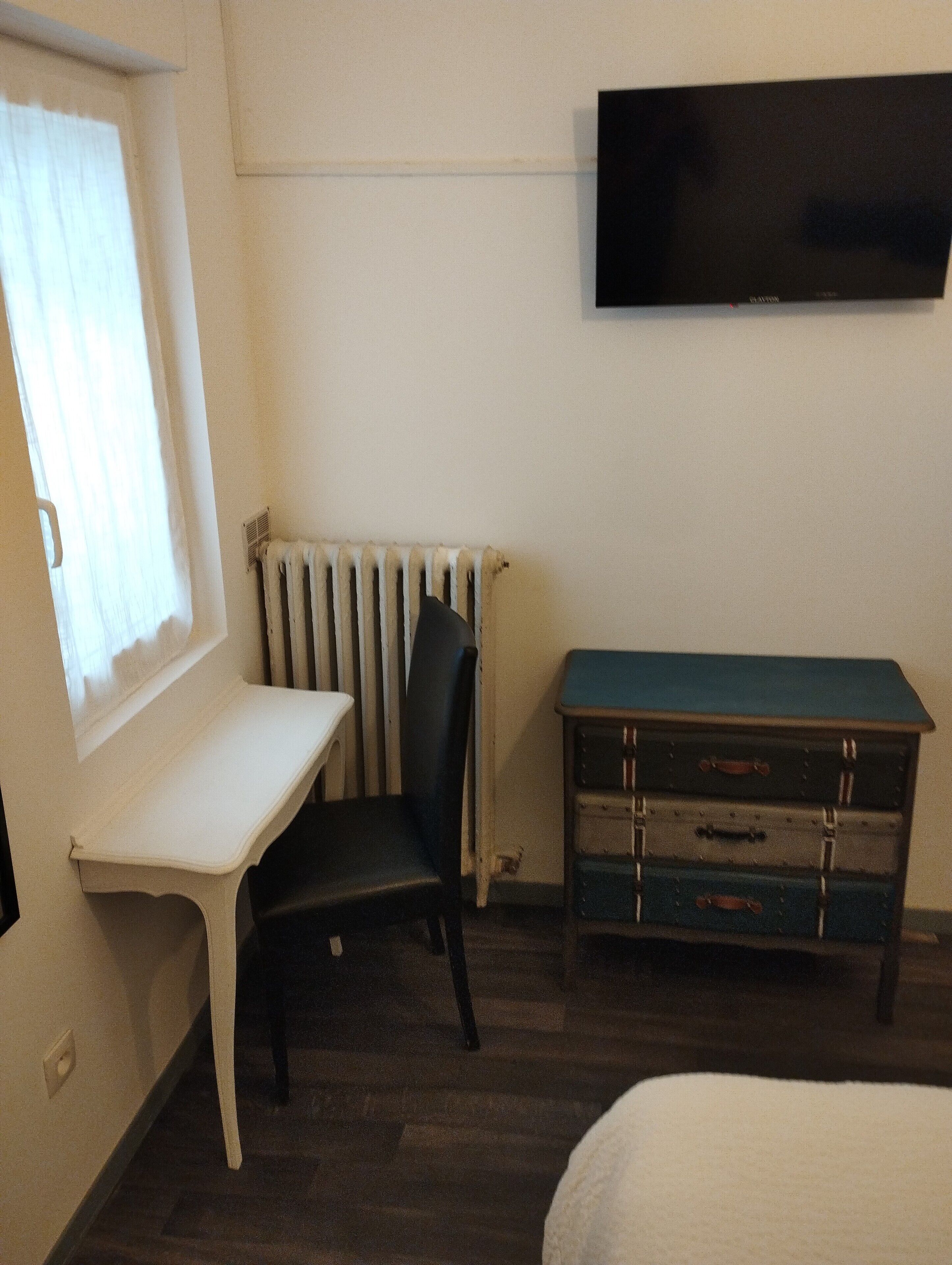 A room with a white desk, a black chair, a dresser with multiple drawers, a radiator, and a mounted television.