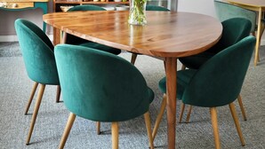 Un comedor con una mesa de madera, sillas tapizadas de verde, un jarrón con flores y estantes con frascos y libros.
