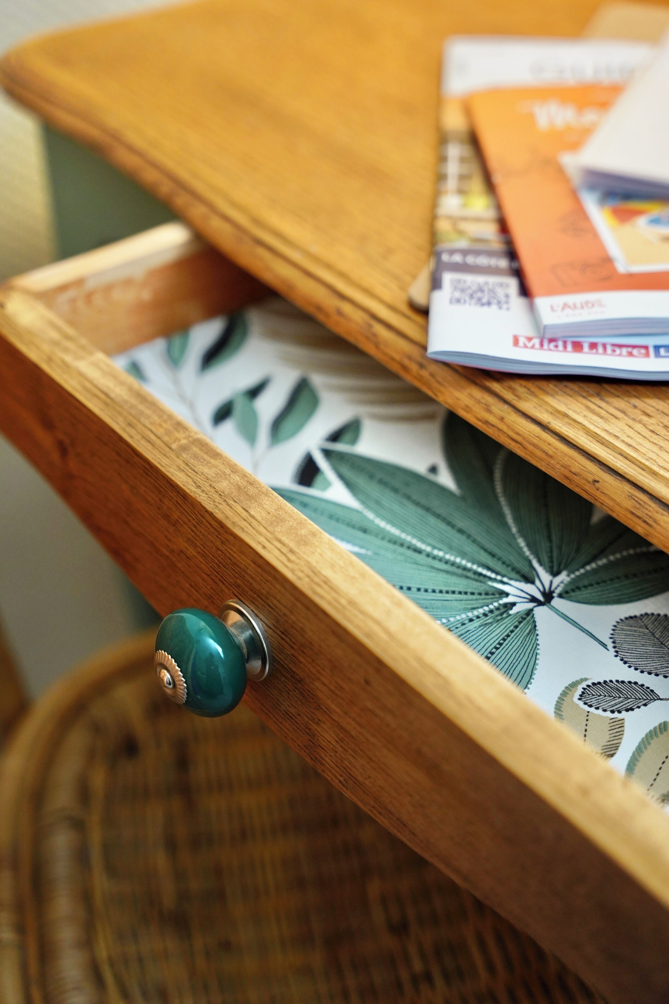 Un cajón de madera con un pomo verde, una cesta de mimbre y una revista encima.