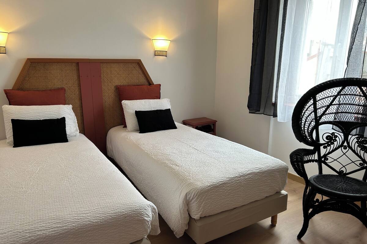 A hotel room with two single beds, a small wooden nightstand, a wicker chair, and a window with curtains.