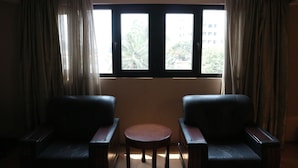 Une pièce avec deux fauteuils en cuir et une petite table ronde, une fenêtre avec des rideaux, et une vue sur les bâtiments et les arbres à l’extérieur.