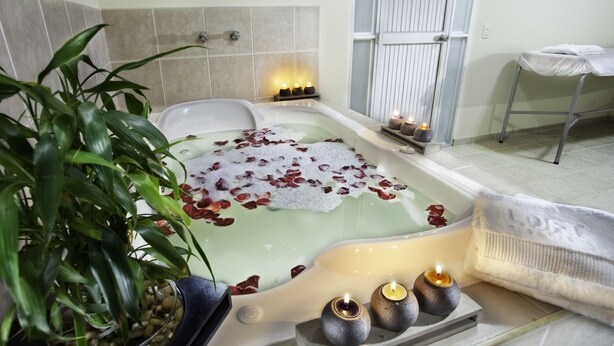 Una bañera con pétalos de rosa, velas y una planta en maceta en un ambiente tipo spa.