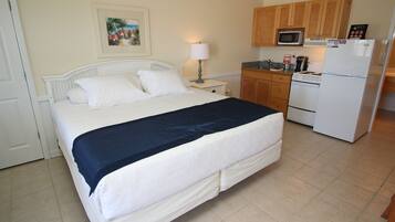 A bedroom with a bed, a nightstand, a lamp, a microwave, a refrigerator, and wooden cabinets.