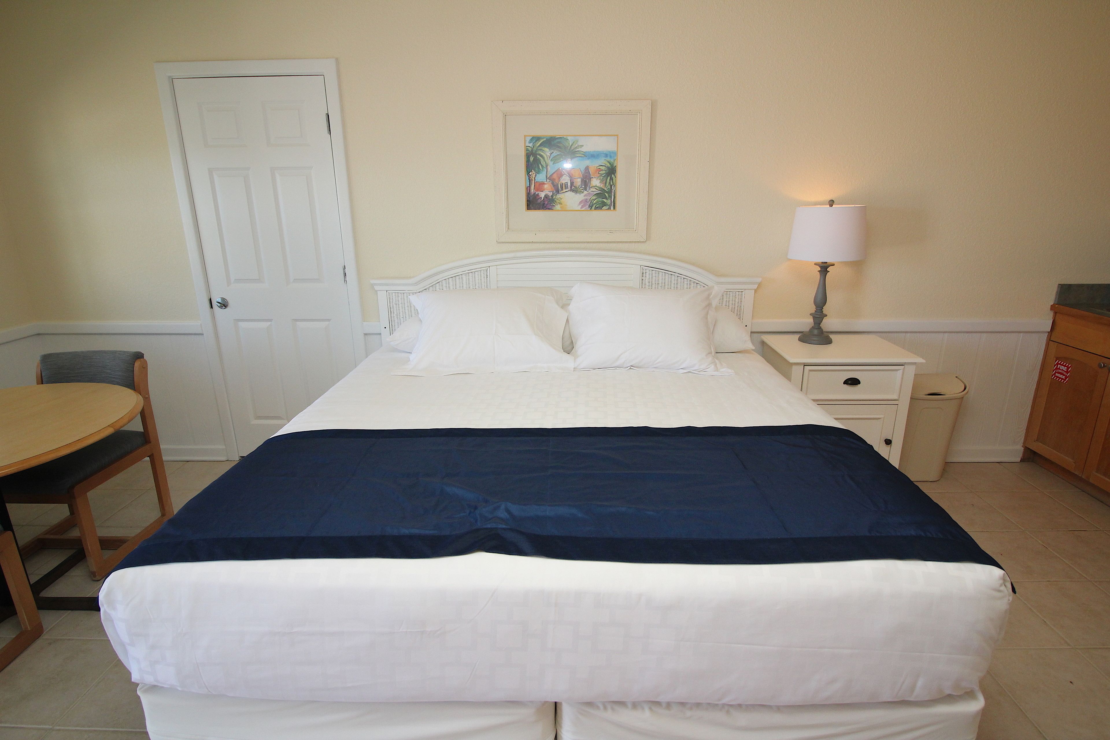 A neatly made bed with white and navy bedding, a bedside table with a lamp, a chair, and a door.