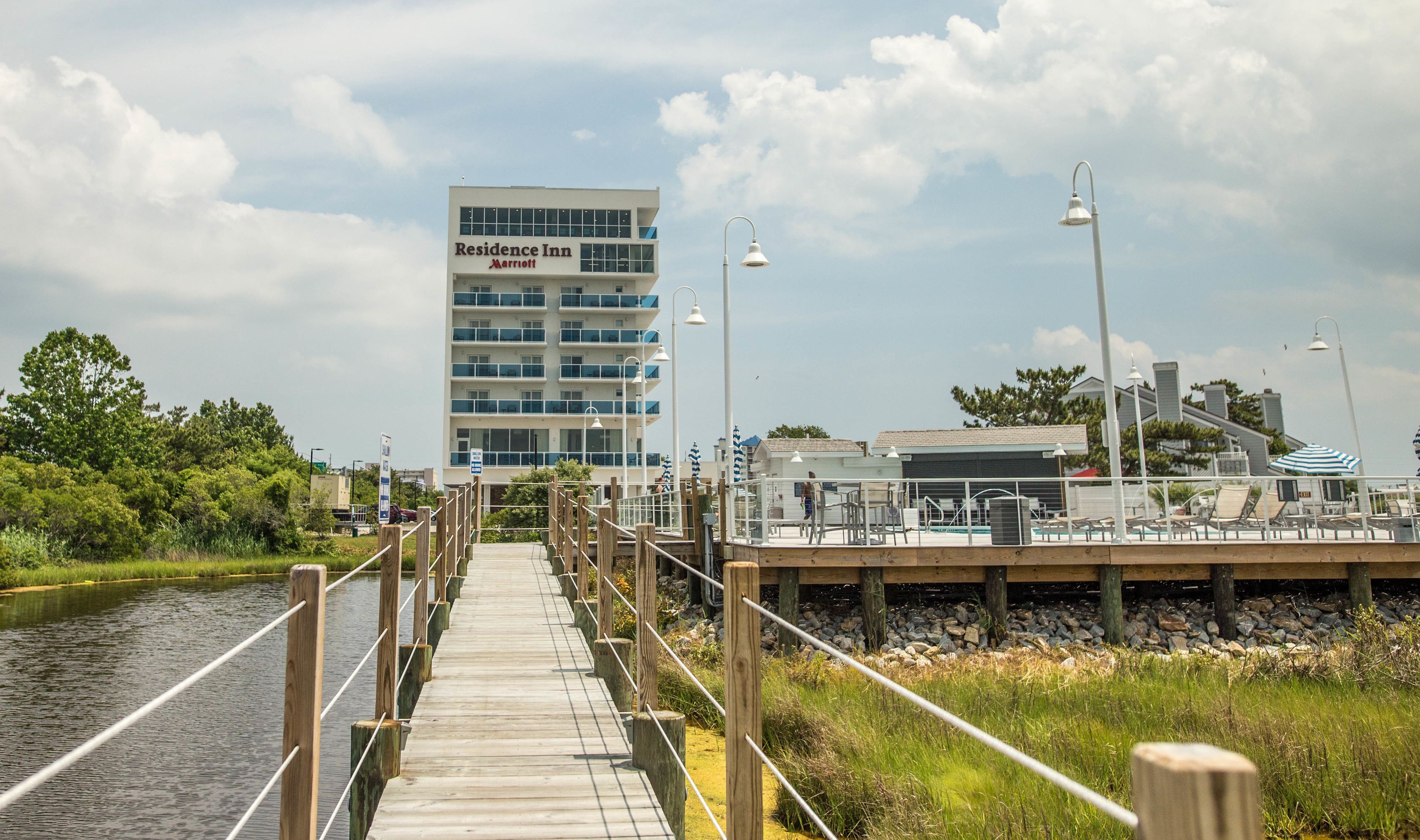 A wooden boardwalk leading to a waterfront area with a Marriott Residence Inn building in the background.