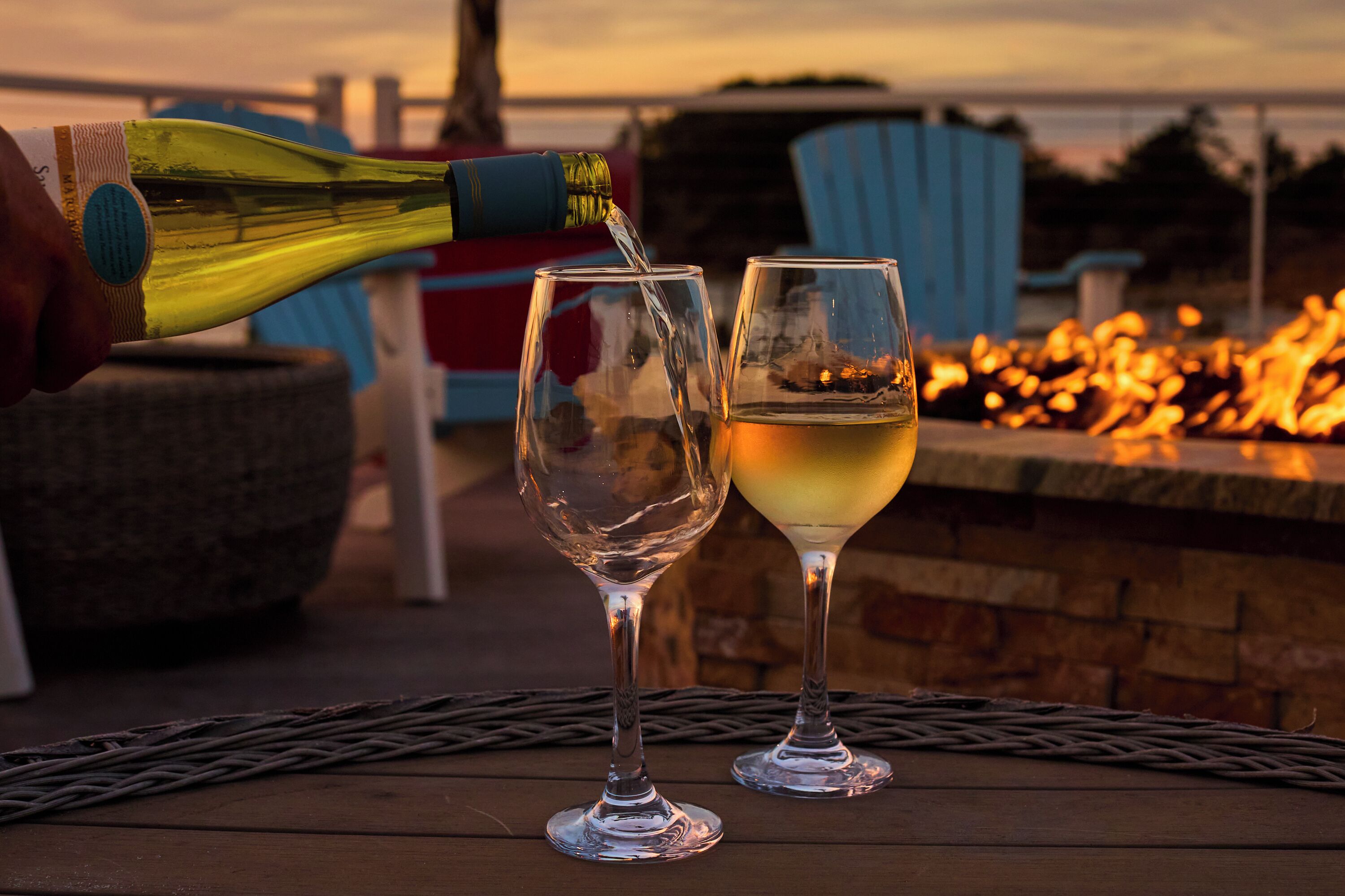 A bottle of white wine being poured into two glasses on a wooden table with a fire pit in the background.