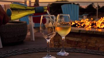 A bottle of white wine being poured into two glasses on a wooden table with a fire pit in the background.