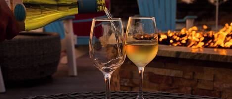 A bottle of white wine being poured into two glasses on a wooden table with a fire pit in the background.