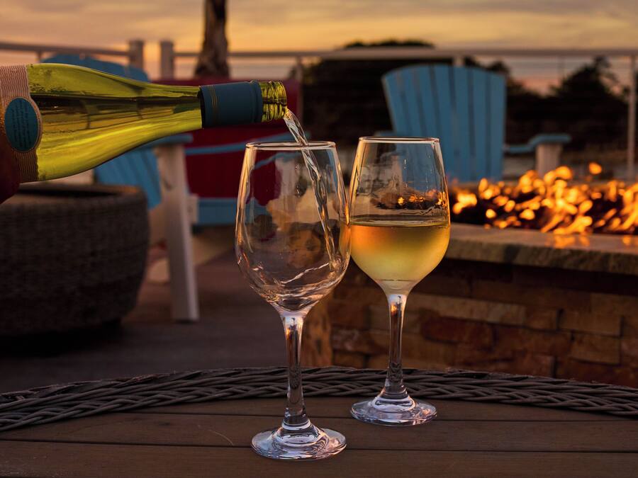 A bottle of white wine being poured into two glasses on a wooden table with a fire pit in the background.