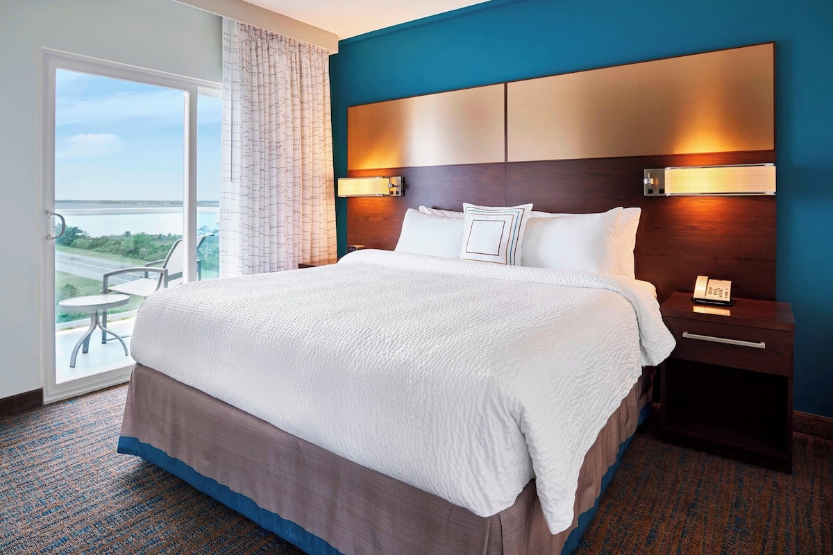 A hotel room with a large bed, a nightstand with a phone, a wall-mounted light, and a view of a balcony with a table and chairs.