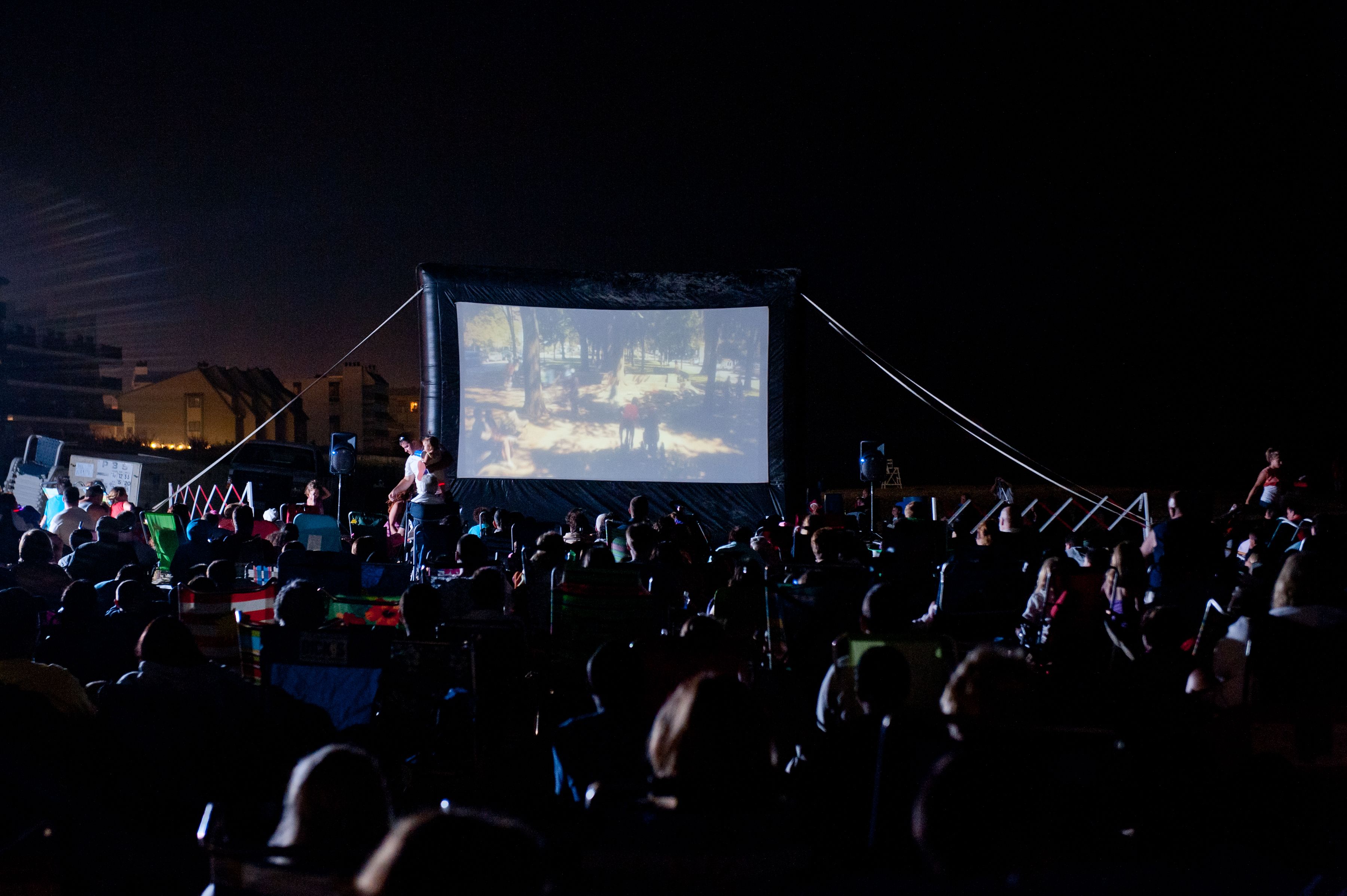 Une projection de film en plein air, avec un grand écran et des spectateurs assis en rangées.