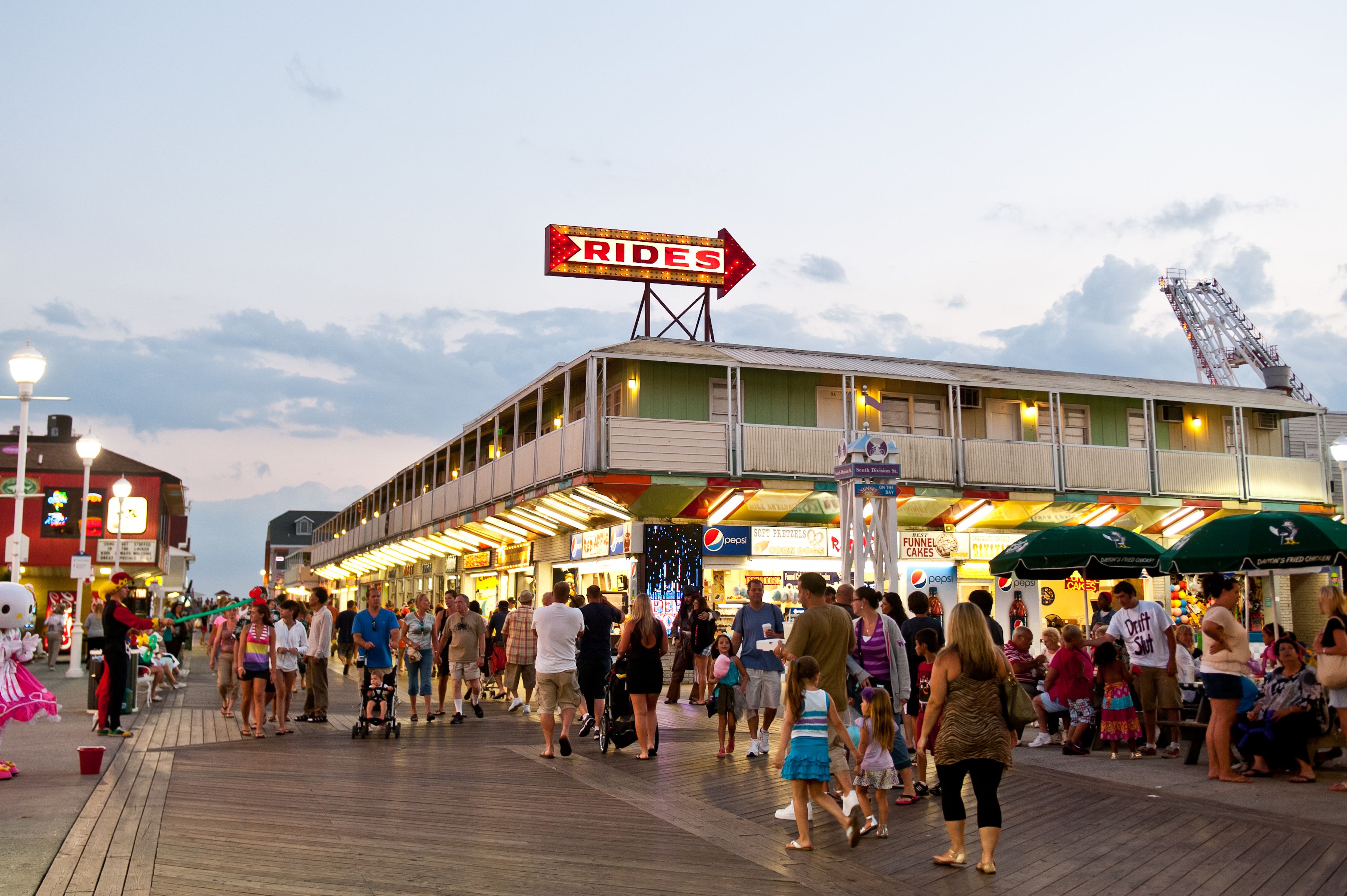 A crowded boardwalk with a sign for 'RIDES' and various shops and food stands.