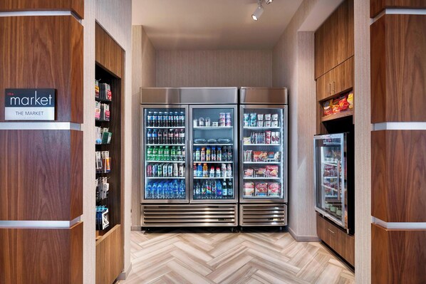 A corridor with wooden walls and a herringbone floor, leading to a refrigerated display case stocked with beverages and snacks.