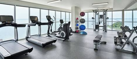 A gym with treadmills, weight machines, and dumbbells, large windows with a view of the sea.
